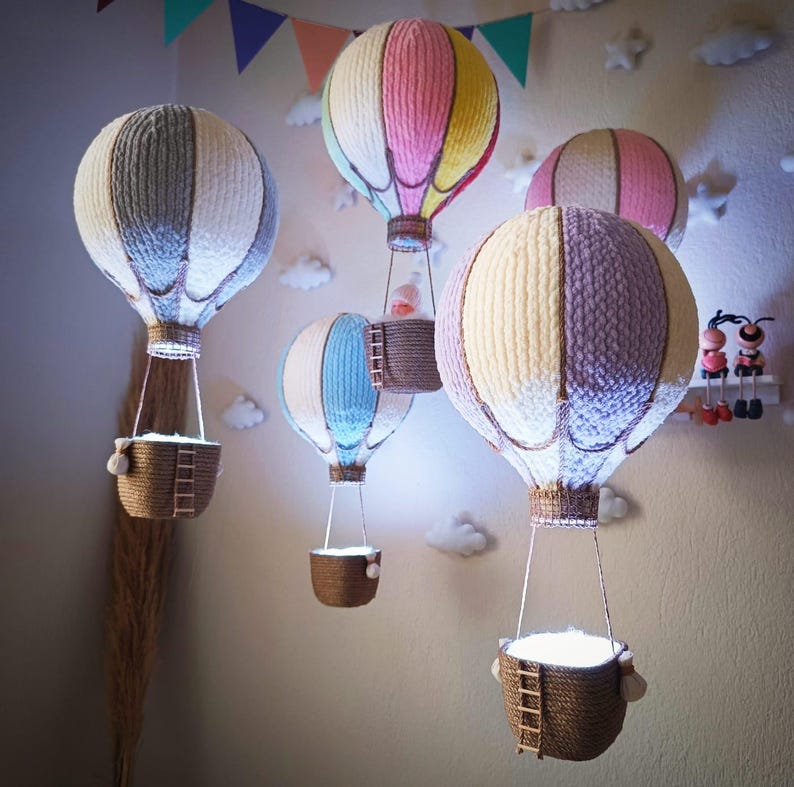 May include: Five colourful hot air balloons with woven fabric and baskets hanging from the ceiling. The balloons are in shades of pink, blue, grey, and white.