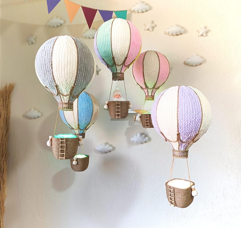 May include: A set of five hot air balloon decorations in various colours, including pink, blue, purple, and white. Each balloon has a woven basket and is suspended from the ceiling.