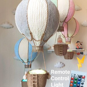 May include: Three white, grey, and pink hot air balloon-shaped lamps with a rope basket and a remote control for changing the light colour. The text "Remote Control Light" is written below the balloons.