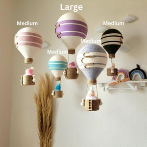 May include: Five handmade hot air balloons in various sizes and colors. The balloons are made of white yarn with colored stripes and have a small basket with a doll inside. The balloons are labeled "Large", "Medium", and "Medium".