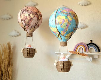 May include: Two decorative hot air balloons with globe designs hang on a wall. One balloon has a pink and brown map design, the other a blue and green world map. Each has a woven basket with a small figure inside.