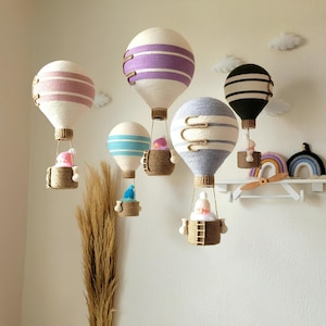 May include: Five white hot air balloons with different colored stripes hang from the ceiling. Each balloon has a small basket with a doll inside. The balloons are decorated with purple, pink, blue, gray, and black stripes.