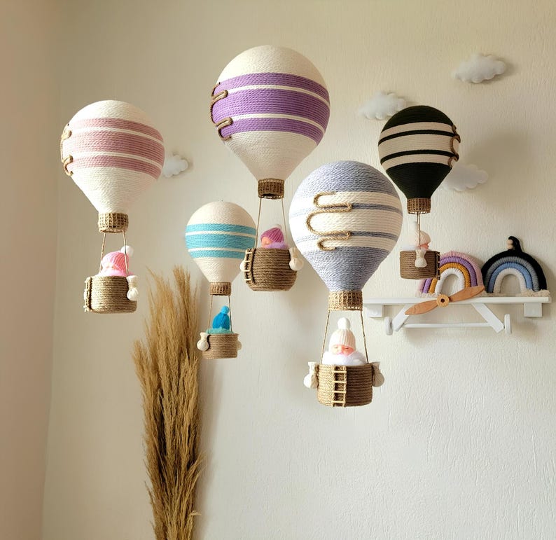 May include: Five white hot air balloons with different colored stripes hang from the ceiling. Each balloon has a small basket with a doll inside. The balloons are decorated with purple, pink, blue, gray, and black stripes.