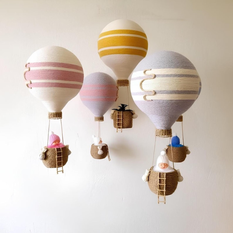 May include: Five decorative hot air balloons made of yarn and fabric, each with a different color scheme and a small basket hanging below. The baskets contain small figures, including a baby doll, a bunny, and a plant.