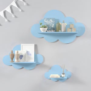 May include: Set of three blue cloud-shaped shelves mounted on a light blue wall. The largest shelf has a book titled "Peter Pan", a small potted plant, a bottle, and wooden blocks that spell "ABC". The middle shelf has two small decorative items and a framed picture. The smallest shelf has a book titled "Wonder War" and a diffuser.
