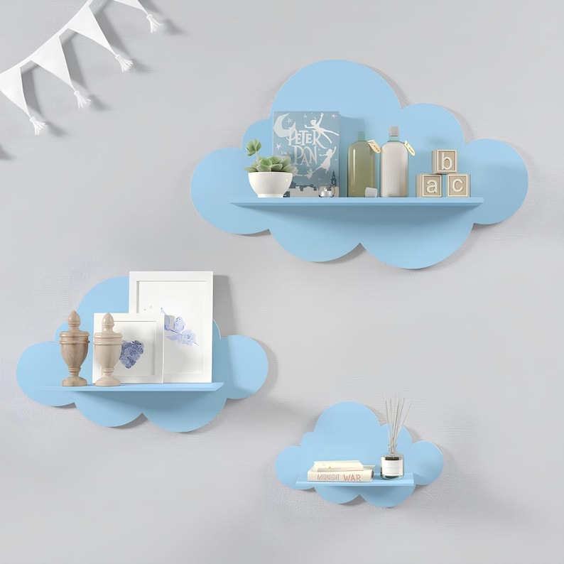 May include: Set of three blue cloud-shaped shelves mounted on a light blue wall. The largest shelf has a book titled "Peter Pan", a small potted plant, a bottle, and wooden blocks that spell "ABC". The middle shelf has two small decorative items and a framed picture. The smallest shelf has a book titled "Wonder War" and a diffuser.