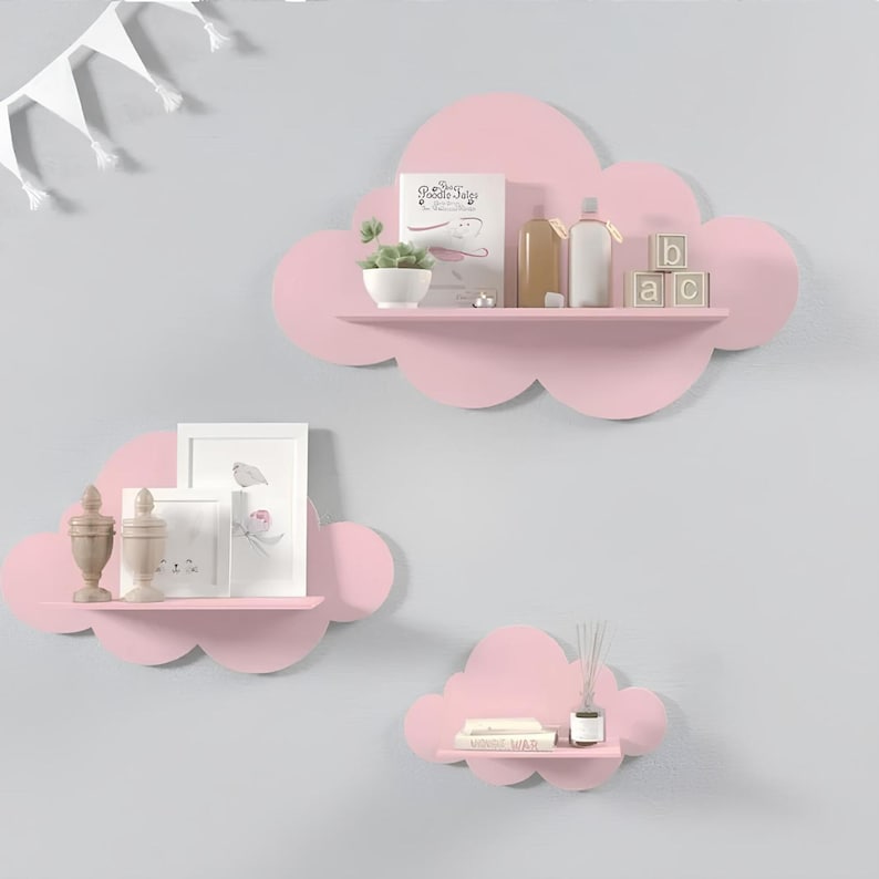 May include: Three pink cloud-shaped shelves mounted on a light gray wall. The top shelf has a small potted plant, two bottles, and three wooden blocks that spell "ABC". The middle shelf has two small decorative items and two framed pictures. The bottom shelf has a book and a diffuser.