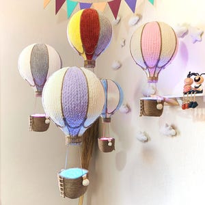 May include: Five colourful hot air balloons with baskets hanging from them. The balloons are made of yarn and are in various colours, including red, yellow, grey, pink, and purple. The baskets are made of woven material and are hanging from the balloons by strings.