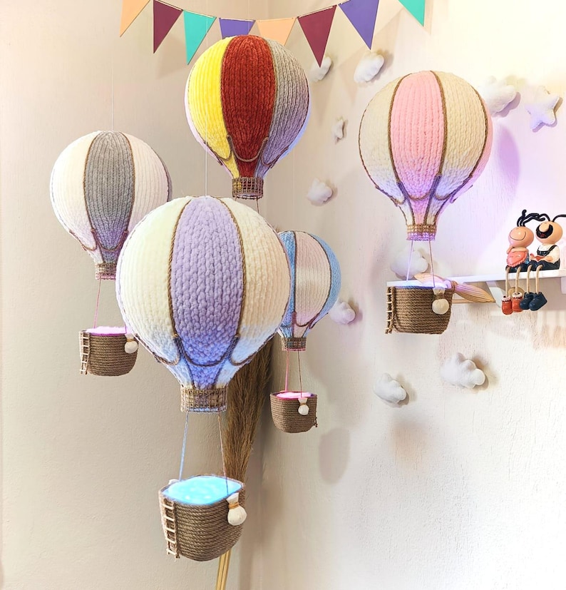 May include: Five colourful hot air balloons with baskets hanging from them. The balloons are made of yarn and are in various colours, including red, yellow, grey, pink, and purple. The baskets are made of woven material and are hanging from the balloons by strings.