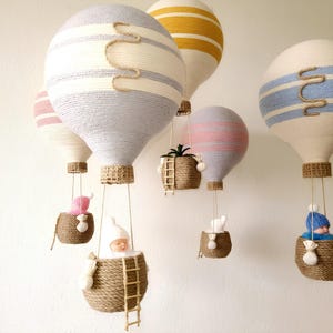 May include: A set of five colorful hot air balloons made of yarn, each with a different pattern and color. The balloons are hanging from the ceiling and each has a small basket underneath. The baskets are filled with various items, including a small doll, a plant, and a small stuffed animal.
