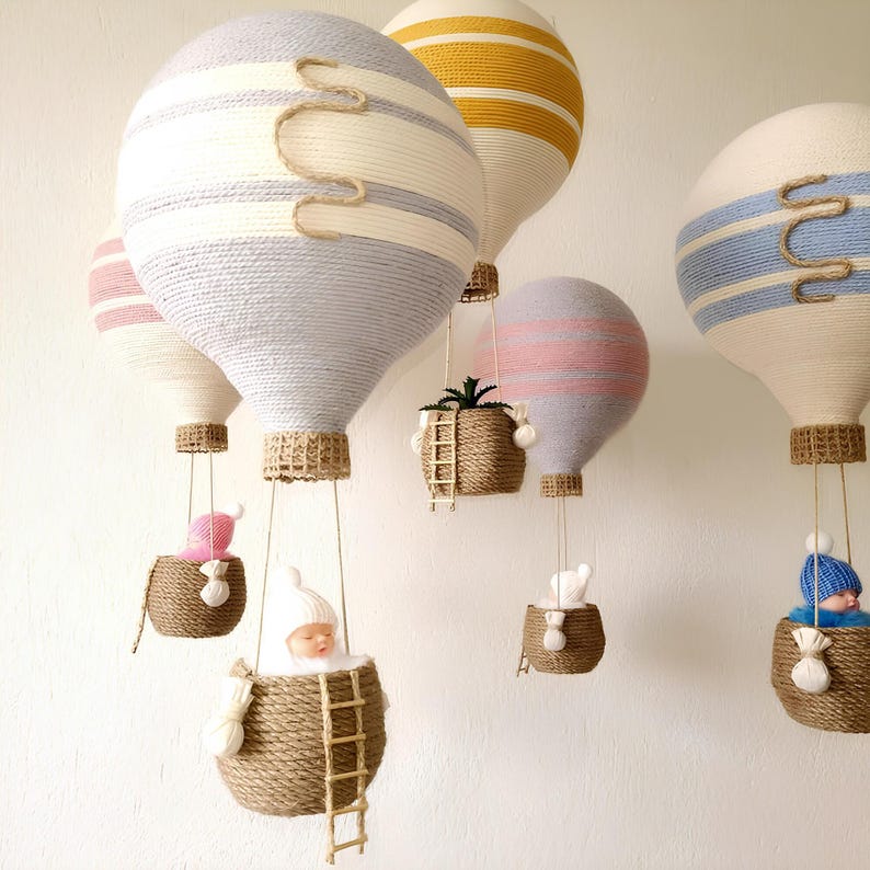 May include: A set of five colorful hot air balloons made of yarn, each with a different pattern and color. The balloons are hanging from the ceiling and each has a small basket underneath. The baskets are filled with various items, including a small doll, a plant, and a small stuffed animal.