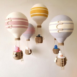 May include: Five white hot air balloons with colorful stripes hang from the ceiling. Each balloon has a small basket with a doll or plant inside. The balloons are made of yarn and have a rustic, handmade look.