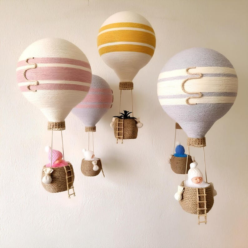 May include: Five white hot air balloons with colorful stripes hang from the ceiling. Each balloon has a small basket with a doll or plant inside. The balloons are made of yarn and have a rustic, handmade look.