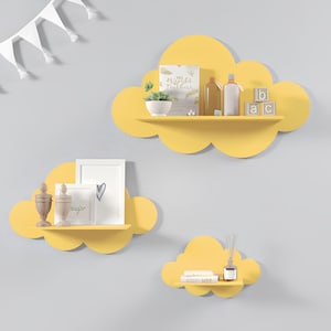 May include: Three yellow cloud-shaped shelves mounted on a gray wall. The largest shelf has a book, a small potted plant, and two bottles on it. The middle shelf has a small picture frame and a decorative vase. The smallest shelf has a book and a diffuser.
