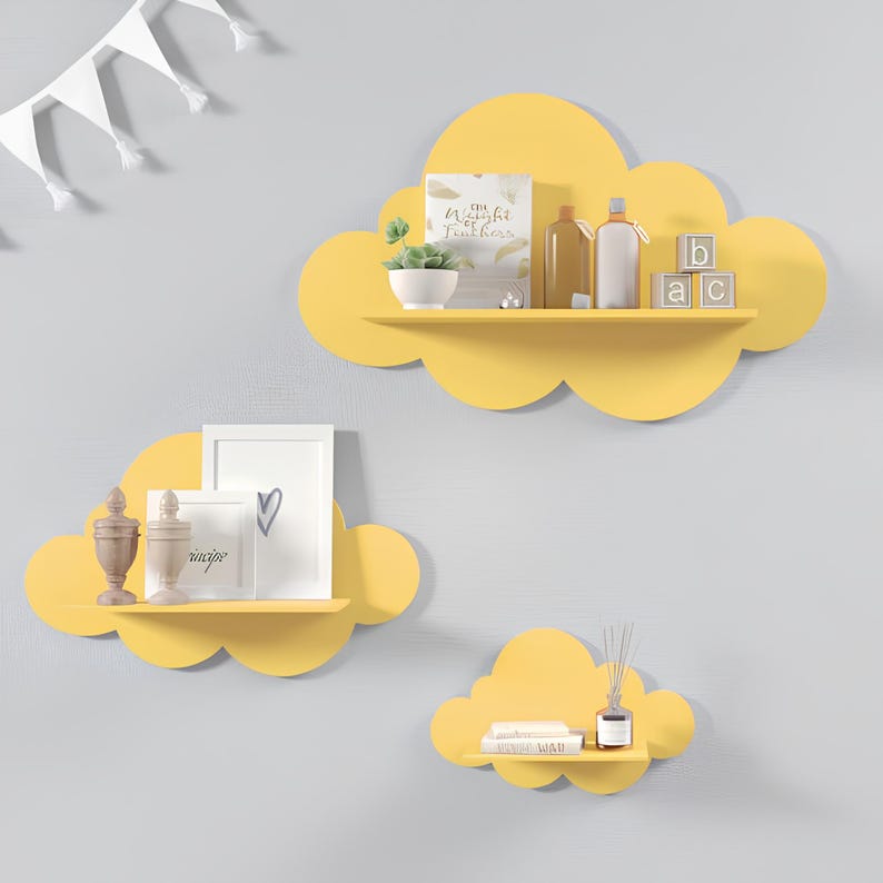 May include: Three yellow cloud-shaped shelves mounted on a gray wall. The largest shelf has a book, a small potted plant, and two bottles on it. The middle shelf has a small picture frame and a decorative vase. The smallest shelf has a book and a diffuser.