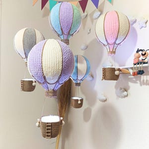 May include: Five colourful hot air balloons with baskets hanging from them. The balloons are made of fabric and have a woven texture. The balloons are in various colours, including pink, purple, blue, and grey. The balloons are hanging from the ceiling and are decorated with string.