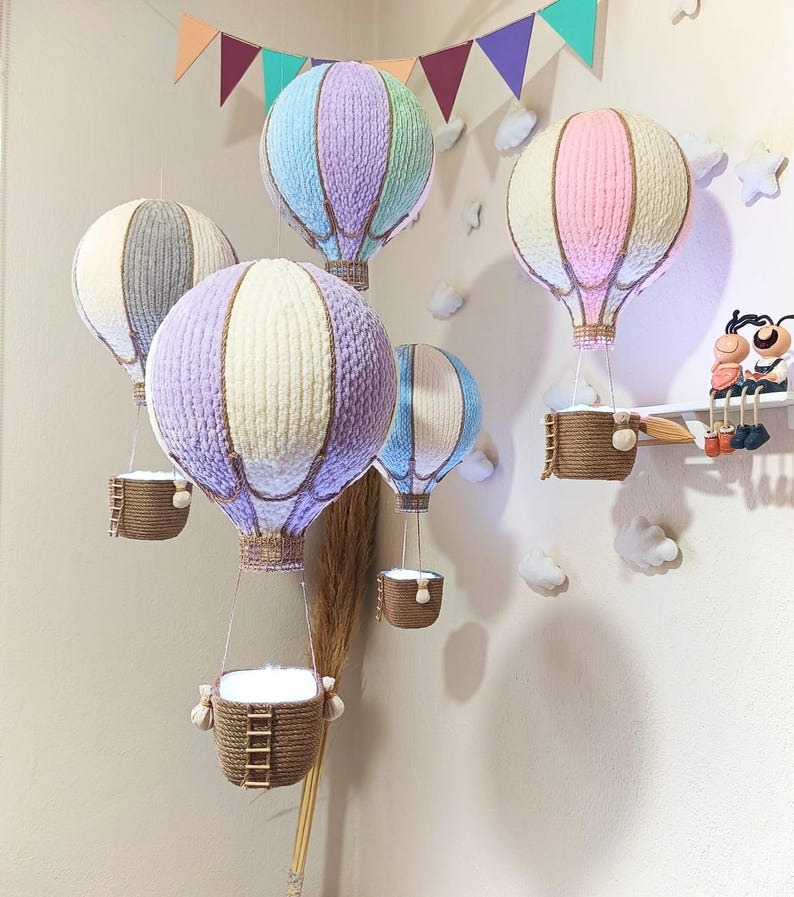 May include: Five colourful hot air balloons with baskets hanging from them. The balloons are made of fabric and have a woven texture. The balloons are in various colours, including pink, purple, blue, and grey. The balloons are hanging from the ceiling and are decorated with string.