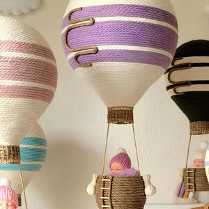 May include: Three hot air balloons made of yarn with a baby doll in each basket. The balloons are decorated with stripes of pink, purple, and blue yarn. The baskets are made of woven brown yarn.