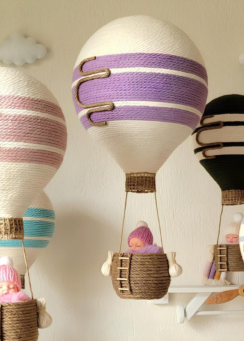 May include: Three hot air balloons made of yarn with a baby doll in each basket. The balloons are decorated with stripes of pink, purple, and blue yarn. The baskets are made of woven brown yarn.