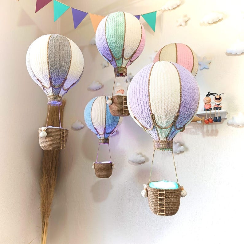May include: Five colourful hot air balloons with woven baskets hanging from the ceiling. The balloons are made of yarn and have a light blue, grey, pink, purple, and green colour scheme.