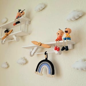 May include: Two white wooden airplane shelves with figurines of people sitting on them. The shelves are mounted on a white wall with white felt clouds. A rainbow macrame hanging is below the shelves.