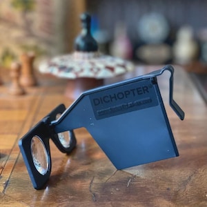 Puede incluir: Gafas dicópticas negras con lentes transparentes y montura rectangular. Las gafas tienen el texto "DICHOPTER®" y "www.SiriusEnterprises.com" impreso en la montura. Las gafas están diseñadas para usarse en la cara.