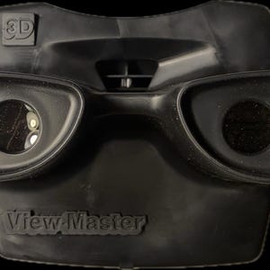 Könnte beinhalten: Ein schwarzer View-Master 3D-Betrachter aus Kunststoff mit zwei runden Betrachtungslinsen. Der Betrachter hat einen schwarzen Kunststoffgriff und der Text "View-Master" ist auf der Vorderseite aufgedruckt.