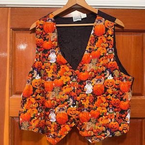 May include: A sleeveless vest featuring a vibrant Halloween-themed print with orange pumpkins, autumn leaves, and small teddy bears dressed in costumes against a black background. The vest has a black interior lining and a button-front closure.