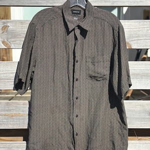 Puede incluir: Camisa de manga corta con estampado de cuadros marrones y negros. La camisa tiene un bolsillo en el pecho y está abotonada. Cuelga de una percha transparente. La tela parece ligera.