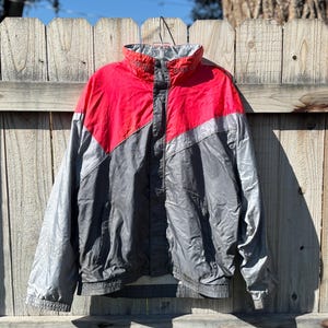 Puede incluir: Chaqueta cortavientos vintage en rojo, gris y plateado. La chaqueta tiene cuello alto y cremallera completa. La palabra "NORTH" es visible en el cuello. La chaqueta está colgada en una valla de madera.