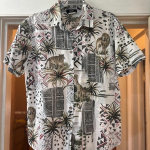 Op de afbeelding: Korte mouwen button-down shirt met een jungle-thema ontwerp op een witte achtergrond. Het patroon bevat leeuwen, tijgers en giraffen, samen met palmbomen en geometrische vormen. Het shirt heeft een kraag.