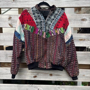 May include: A colorful, patterned bomber jacket with a black zipper and cuffs. The jacket features a patchwork design with red, blue, green, and purple accents. The sleeves and body are a brown plaid. The jacket is hanging on a black hanger.