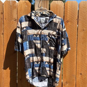 May include: Short-sleeved button-down shirt with a geometric pattern in black, blue, and beige. The shirt has a collared neckline and a single chest pocket. The fabric appears to be lightweight, suitable for warm weather.