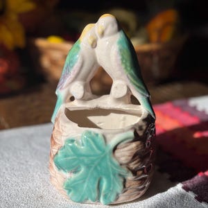 May include: A ceramic planter featuring two kissing birds with white bodies, teal wings, and yellow beaks. The planter has a brown, textured base with a teal leaf accent. The planter is approximately 4 inches tall.