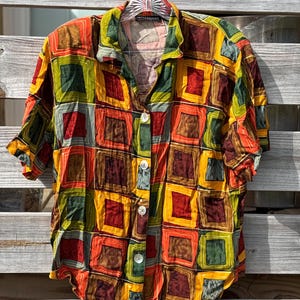 May include: A short-sleeved button-down shirt with a colorful geometric pattern. The shirt features a repeating design of squares in shades of red, orange, yellow, green, and brown. The shirt has a collar and three white buttons. The brand name "IMPRESSIONE" is visible.