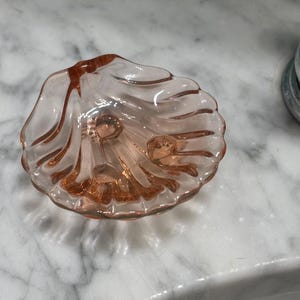 May include: A small, pink glass dish shaped like a seashell. The dish has a scalloped edge and a textured, ridged design. The glass is translucent, allowing light to pass through. A chrome fixture is visible in the background.