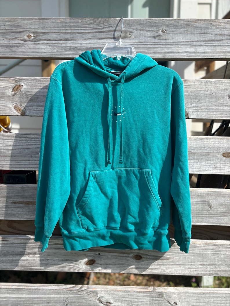 May include: Teal hoodie with a front pocket and a drawstring hood. The hoodie has a small white logo on the chest. The garment is hanging on a clear plastic hanger against a wooden fence.