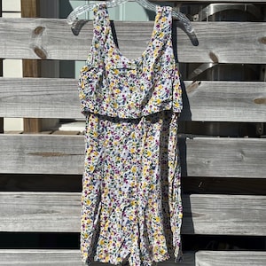 May include: A sleeveless romper with a floral pattern in white, yellow, purple, and blue. The romper has a ruffled top and is hanging on a clear plastic hanger against a wooden fence.