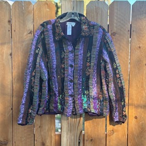 May include: A multi-colored jacket with a striped pattern in shades of purple, black, and brown. The jacket has a button-up front, a collar, and floral embroidery. The jacket is hanging on a wooden fence.
