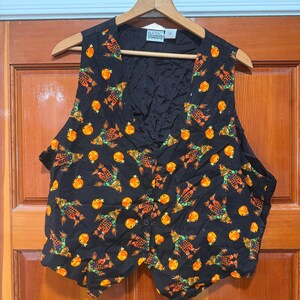 May include: A black sleeveless vest featuring a repeating pattern of orange pumpkins and scarecrows. The vest has a V-neckline, a front button closure with four black buttons, and a label at the inner neckline that reads Basic Editions.