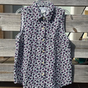 Può includere: Camicia senza maniche con motivo floreale bianco di piccoli fiori viola e verdi. La camicia ha un colletto ed è abbottonata sul davanti. Il testo "WET SEAL" è visibile sul colletto. È appesa a una gruccia di plastica trasparente.