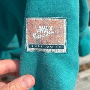 May include: Teal sweatshirt sleeve with a distressed Nike logo patch. The patch features the Nike swoosh and the words "JUST DO IT" in white text. The fabric appears soft and worn.