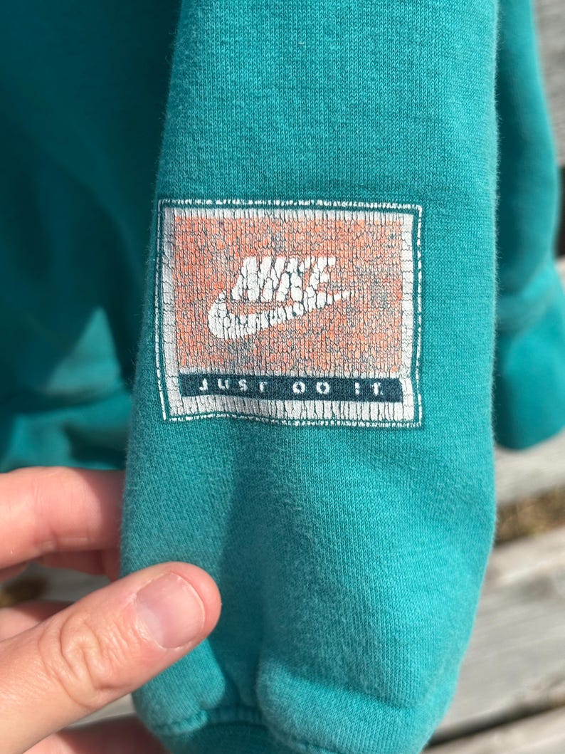 May include: Teal sweatshirt sleeve with a distressed Nike logo patch. The patch features the Nike swoosh and the words "JUST DO IT" in white text. The fabric appears soft and worn.