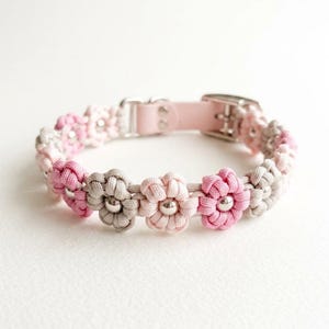Pode incluir: Uma coleira com uma série de flores de cordão trançado em tons alternados de rosa claro, rosa escuro e cinza. Cada flor tem uma pequena conta de metal prateado no centro. A coleira inclui uma seção de tira rosa claro com uma fivela de metal.