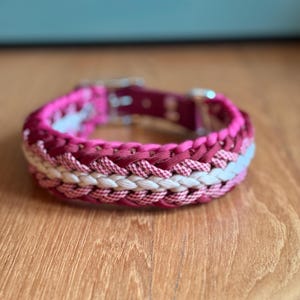 Può includere: Un collare intrecciato per cani nei toni del rosa e del bianco, con una fibbia in metallo color argento. Il collare è realizzato in materiale tessuto e poggia su una superficie di legno.