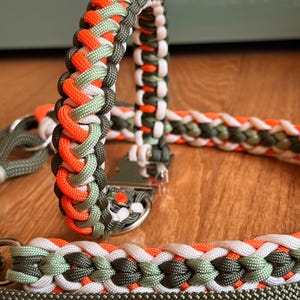 Op de afbeelding: Close-up van een halsband en riem voor honden. De halsband en riem zijn gemaakt van gevlochten nylon touw in oranje, groen, wit en donkergroen. De halsband heeft een metalen sluiting. De riem heeft een metalen ring.