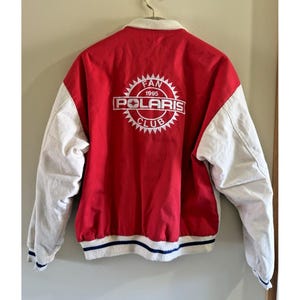 Puede incluir: Una chaqueta de estilo universitario roja y blanca con cuello y mangas largas blancas. La parte posterior muestra un gráfico circular blanco con el texto "FAN 1995 POLARIS CLUB" y un icono de estrella. El dobladillo y los puños son blancos con rayas azul marino.