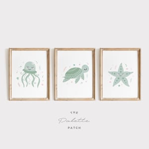 Könnte beinhalten: Drei gerahmte Kunstdrucke mit Meeresbewohnern. Die Drucke zeigen eine lächelnde Qualle, eine Schildkröte und einen Seestern, alle in sanften Grüntönen. Die Rahmen sind aus hellem Holz, was eine sanfte und einladende Ästhetik schafft.