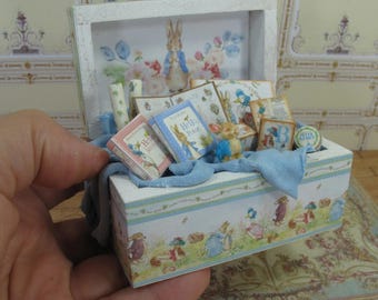 Könnte beinhalten: Eine weiße Holzkiste mit einem blauen und grünen Blumenmuster. Die Kiste ist gefüllt mit Miniaturbüchern, Spielzeug und anderen Gegenständen. Die Kiste ist mit einem Peter Rabbit-Motiv verziert.