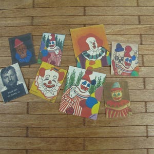 Peut inclure: Neuf cartes colorées illustrant des clowns. Les cartes sont sur une surface en bois. Une carte présente un portrait-robot d'une personne avec le texte "Police Dept. Big Plains, IL."